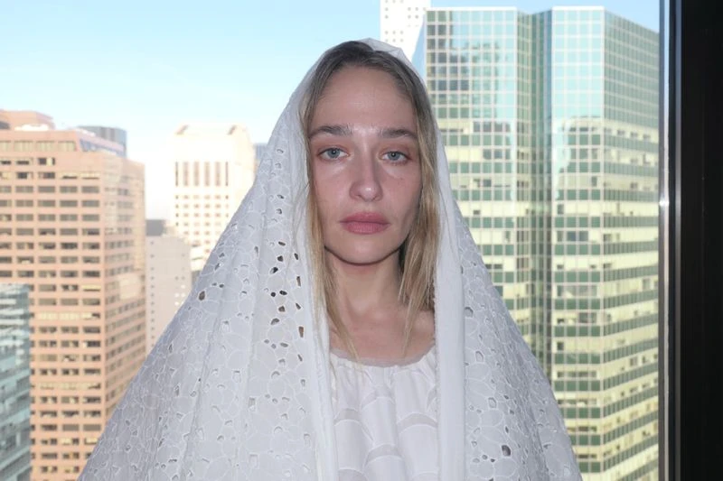 Woman with blond hair poses in front of a city skyline, wearing a white lace veil and a white dress, in a medium shot.