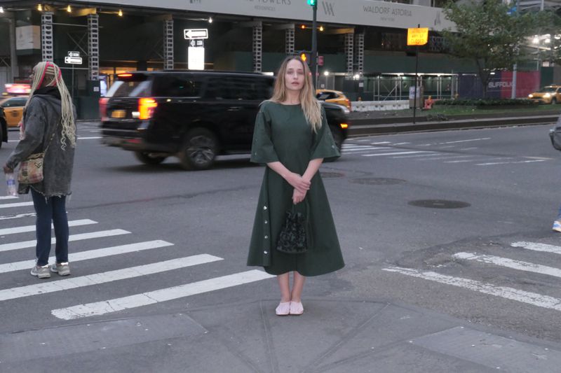 Woman in green dress stands on New York City crosswalk, holding a small bag, with city buildings and traffic in the background.