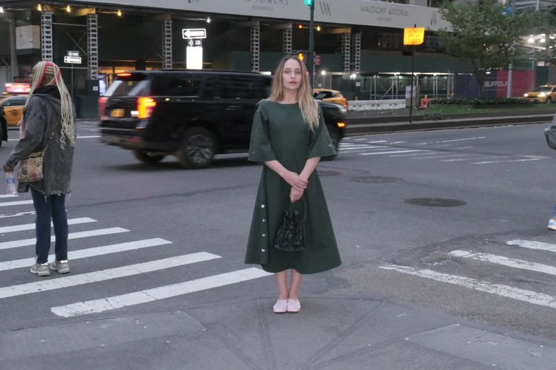 Woman in green dress stands on New York City crosswalk, holding a small bag, with city buildings and traffic in the background.