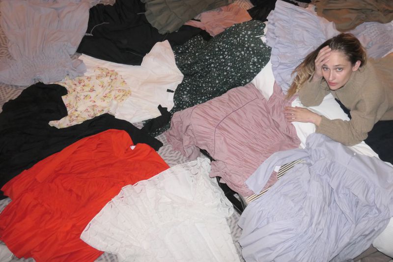 Adult woman lying on patterned blanket, surrounded by piles of colourful and neutral-toned shirts and dresses in an indoor setting.
