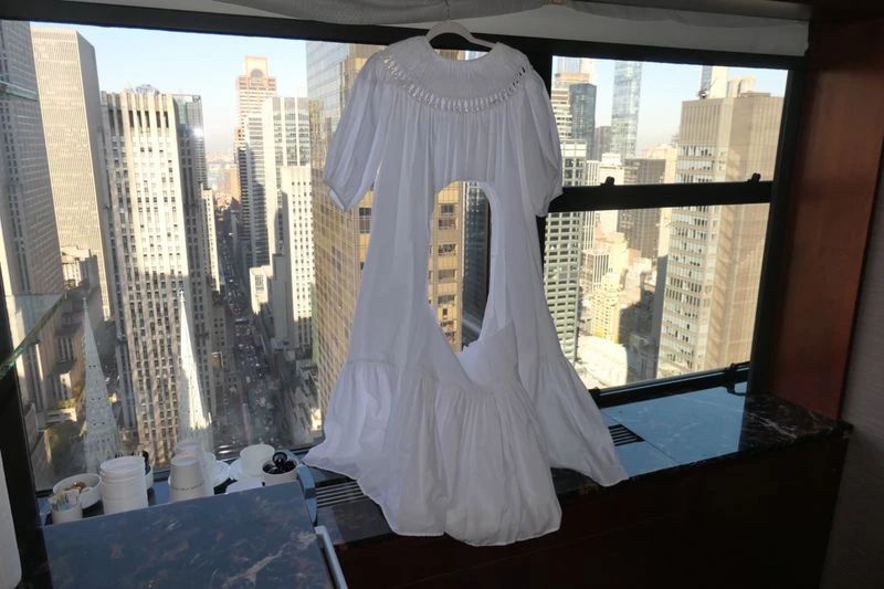 White dress hangs on a hanger in front of a window with city views. Natural light illuminates the garment and the cityscape.
