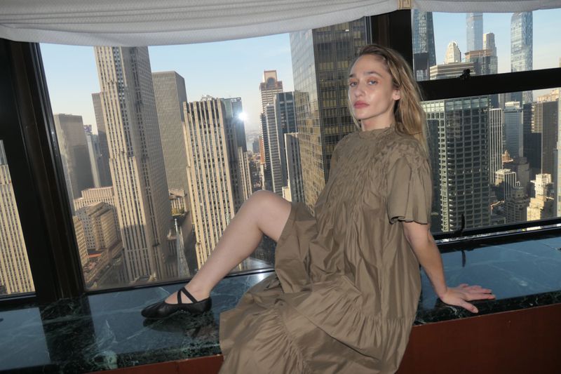 Young woman in a loose brown dress and ballet shoes poses on a marble ledge in front of a window overlooking a city skyline.