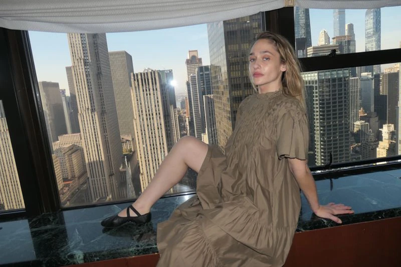 Young woman in a loose brown dress and ballet shoes poses on a marble ledge in front of a window overlooking a city skyline.