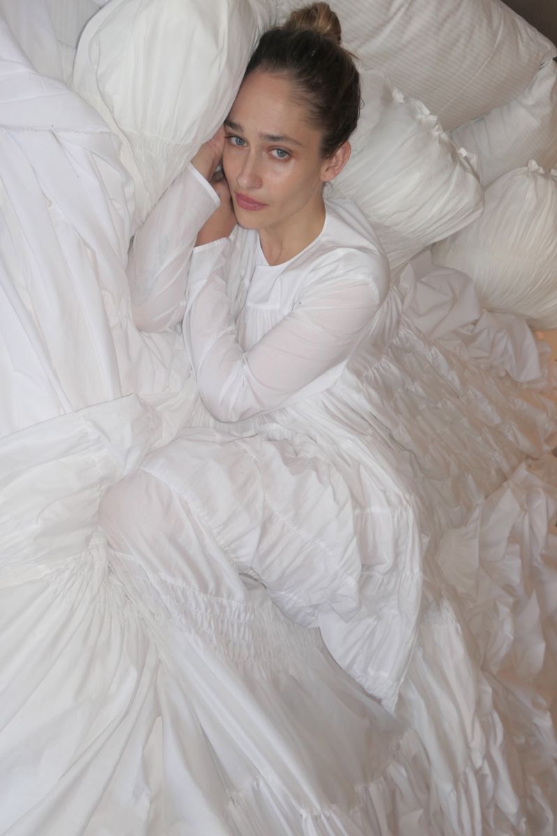 Woman in a white dress lies on a pile of soft white cushions, resting her head on her hand, looking at the camera with a serene expression.