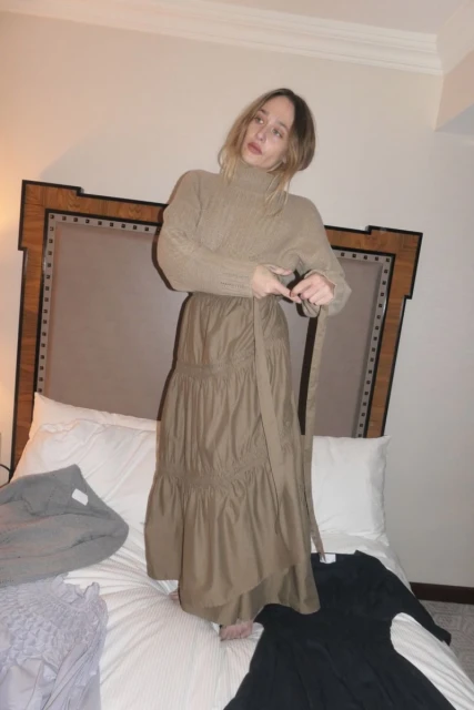 Woman in a tan sweater and flowing brown skirt ties a belt on a bed in an indoor setting with soft, diffused light.