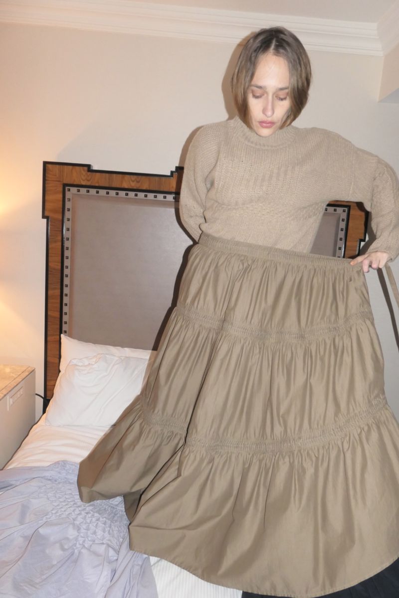 Adult woman poses indoors in a tan cable knit sweater and a tiered skirt. A bed and headboard appear in the background.