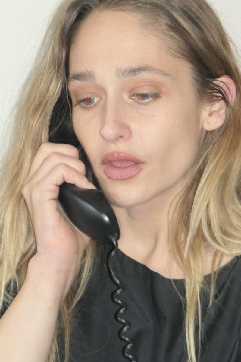 Close-up of a blonde woman in a black top talking on a black landline telephone with a coiled cord against a white backdrop.