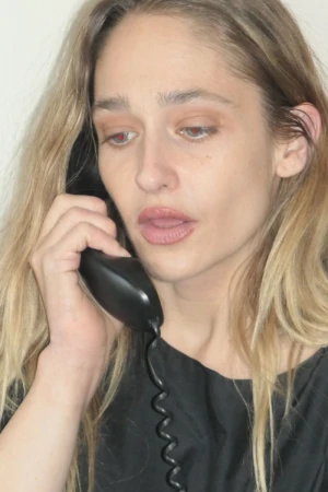 Close-up of a blonde woman in a black top talking on a black landline telephone with a coiled cord against a white backdrop.