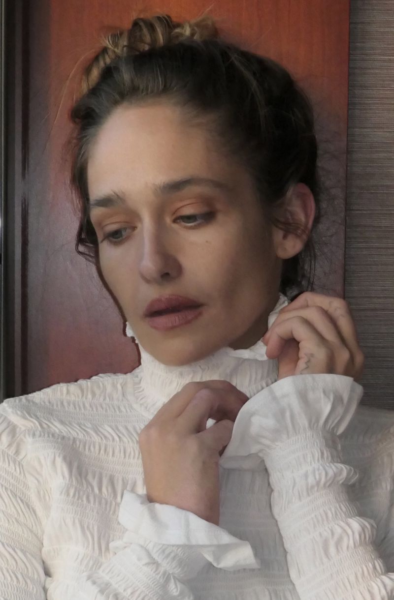 Portrait of an adult woman with hair pulled back wearing a high-collared textured white blouse, adjusting the collar in an indoor setting.
