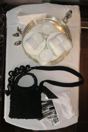 Overhead shot of black handbag, silverware, dishes, salt and pepper shakers, and white linens on a white table.