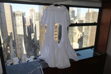 White dress hanging on a hanger in front of a window overlooking a city skyline with skyscrapers.