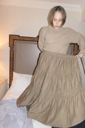 Model Jemima Kirke wearing a tan knit sweater and tiered skirt in a bedroom interior for Merlette Pre Fall 2025 Campaign