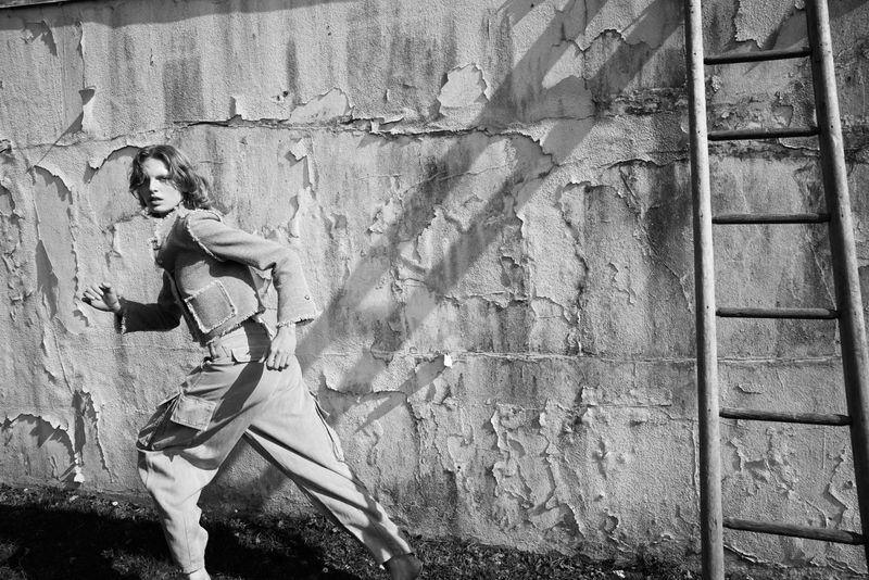 Monochrome portrait of a woman in jacket and pants, moving in front of a textured wall with a ladder, artistic fashion photography.