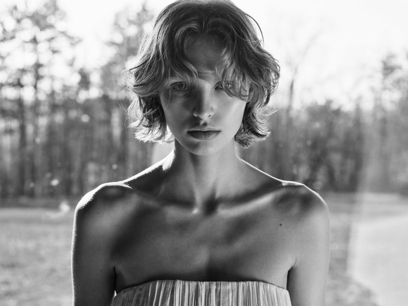 Black and white portrait of a young adult woman with short wavy hair and a strapless dress against a blurred background of trees.
