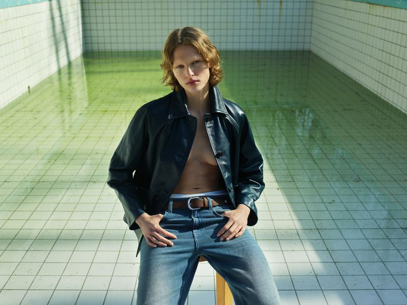 Nonbinary person in leather jacket and jeans sits in drained pool with tiled walls. Moody fashion shot of androgynous individual.