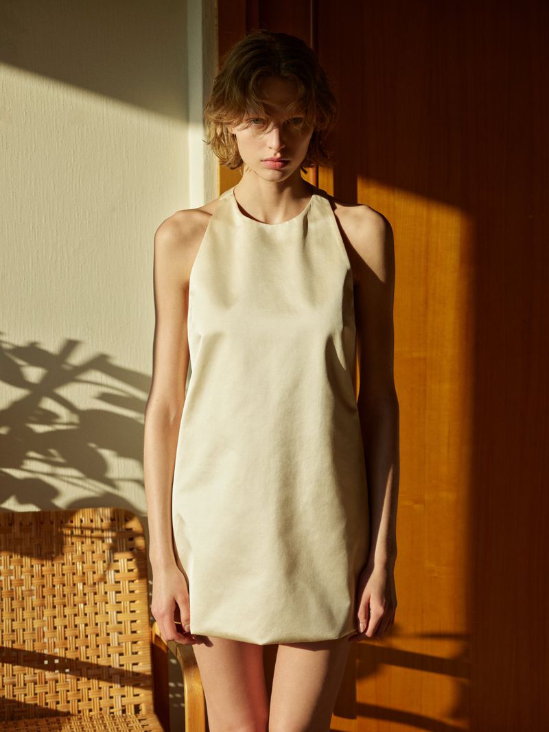 Woman wearing cream-colored halter dress standing indoors in sunlight, posed near a woven chair and wooden door. Fashion editorial style.