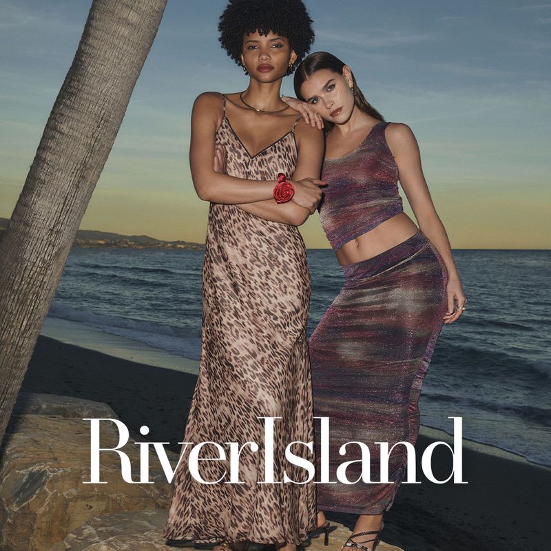 Two fashion models pose on a rocky outcrop overlooking the ocean at dusk, wearing dresses by River Island; one model wears an animal print dress.