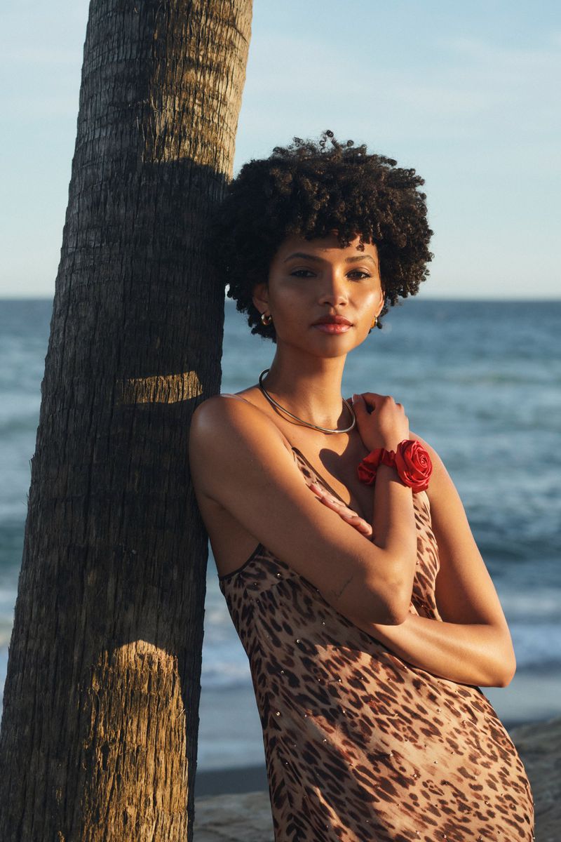 A woman with an afro, wearing a leopard-print dress and a flower bracelet, leans against a palm tree with the ocean and sky in the background.