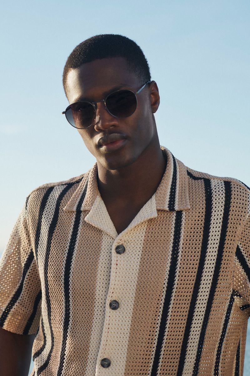 Close-up of a dark-skinned male model wearing round sunglasses and a vertical-striped button-down shirt against a blue sky backdrop.