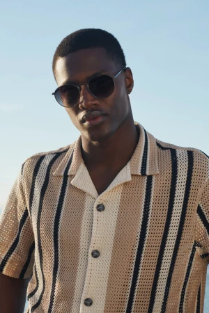 Close-up of a dark-skinned male model wearing round sunglasses and a vertical-striped button-down shirt against a blue sky backdrop.