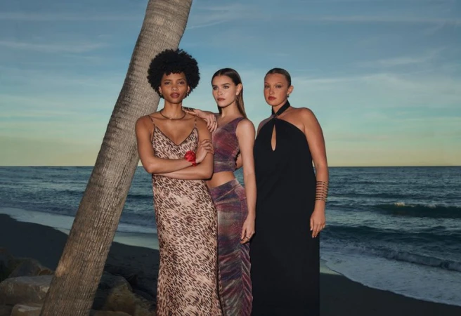 Three fashion models in evening gowns pose near a palm tree against an ocean backdrop at dusk, exuding elegance and style.