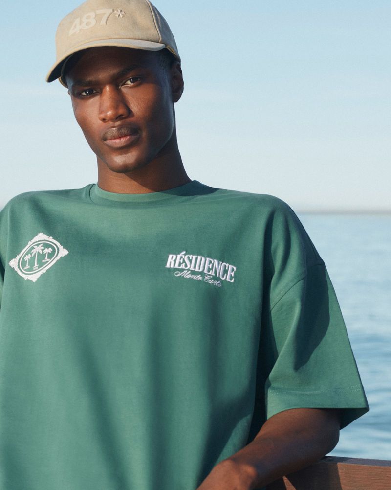 An adult male wearing a green t-shirt and tan baseball cap poses outdoors by the ocean against a light blue sky on a sunny day.