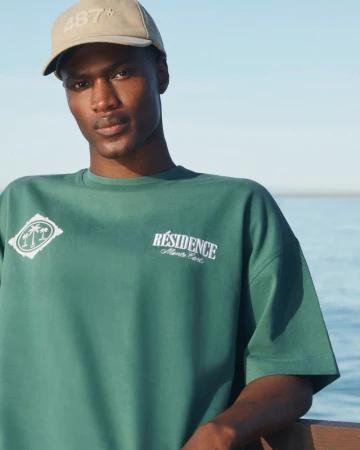 An adult male wearing a green t-shirt and tan baseball cap poses outdoors by the ocean against a light blue sky on a sunny day.