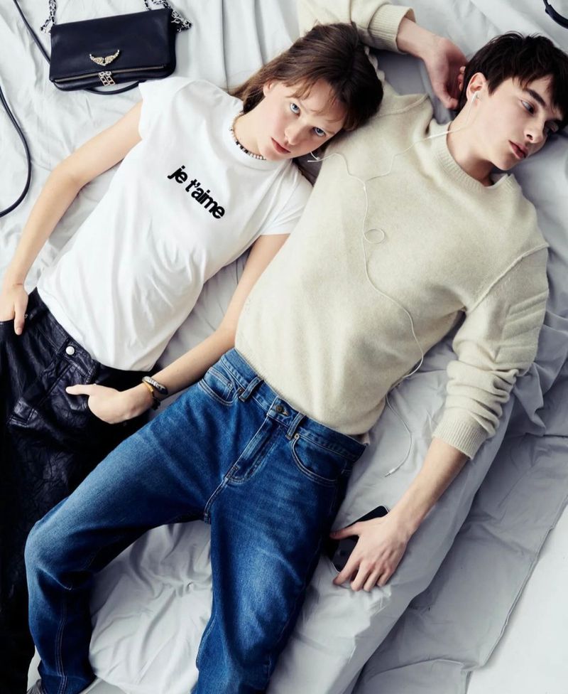 Two young adults pose on a bed with blue jeans, sweaters, and a shoulder bag in a casual, lifestyle-oriented fashion shot.