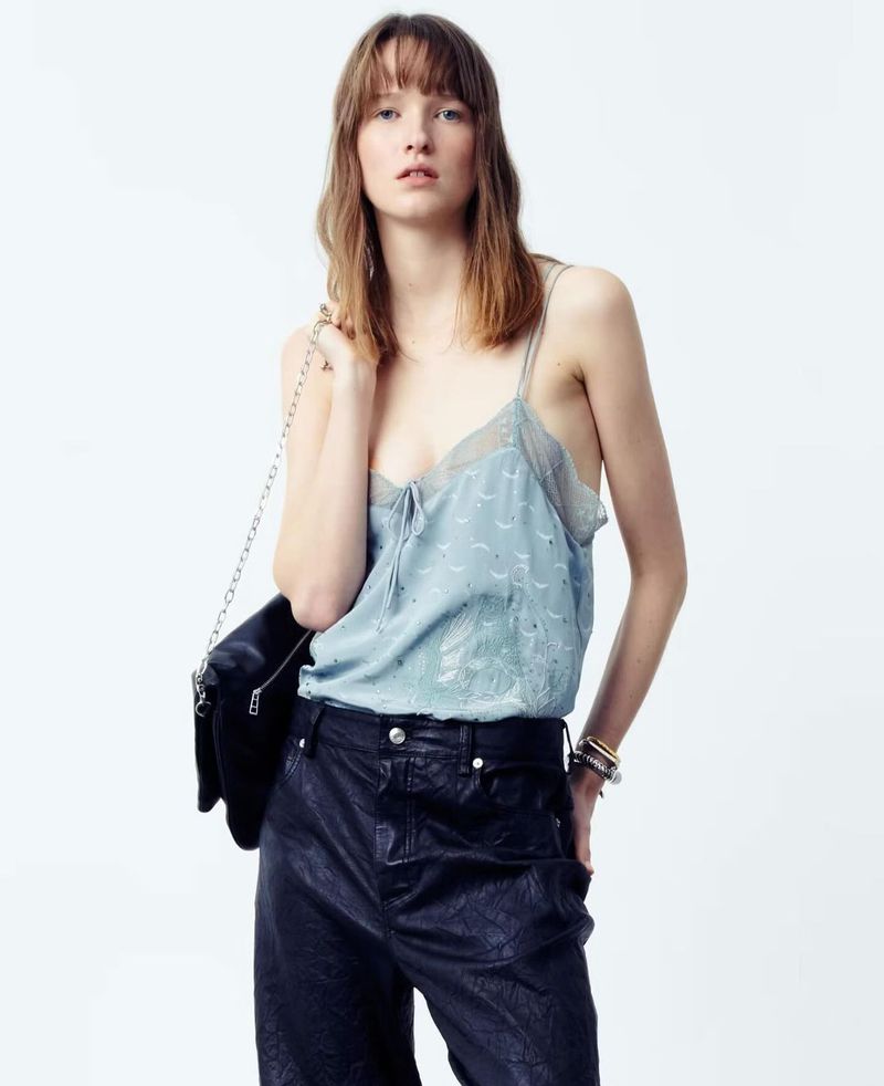 Indoor studio shot of a young adult woman wearing a pale blue tank top, black pants, and a black bag over her shoulder.