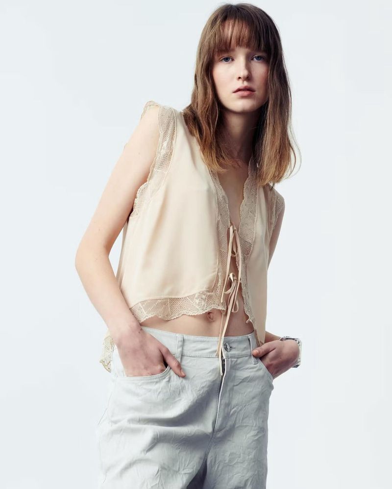 A young woman poses indoors, wearing a lace-trimmed top and light-colored pants, with hands casually in pockets against a bright white backdrop.