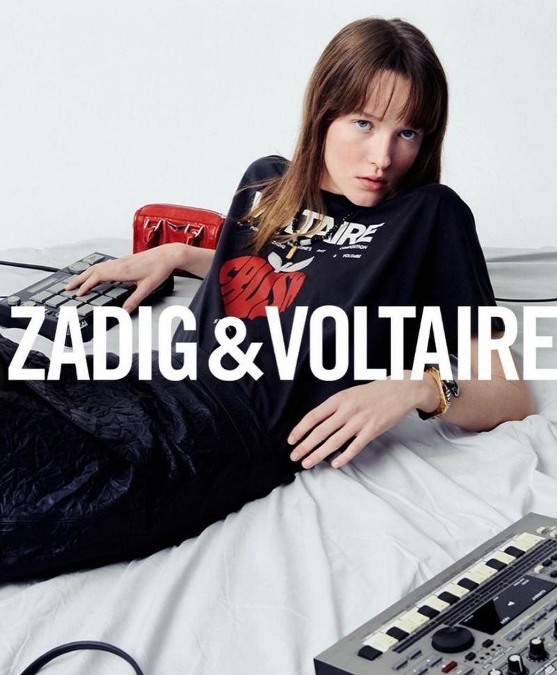 A young woman poses in a black graphic tee on a white backdrop, surrounded by music keyboards and a red patent-leather purse.