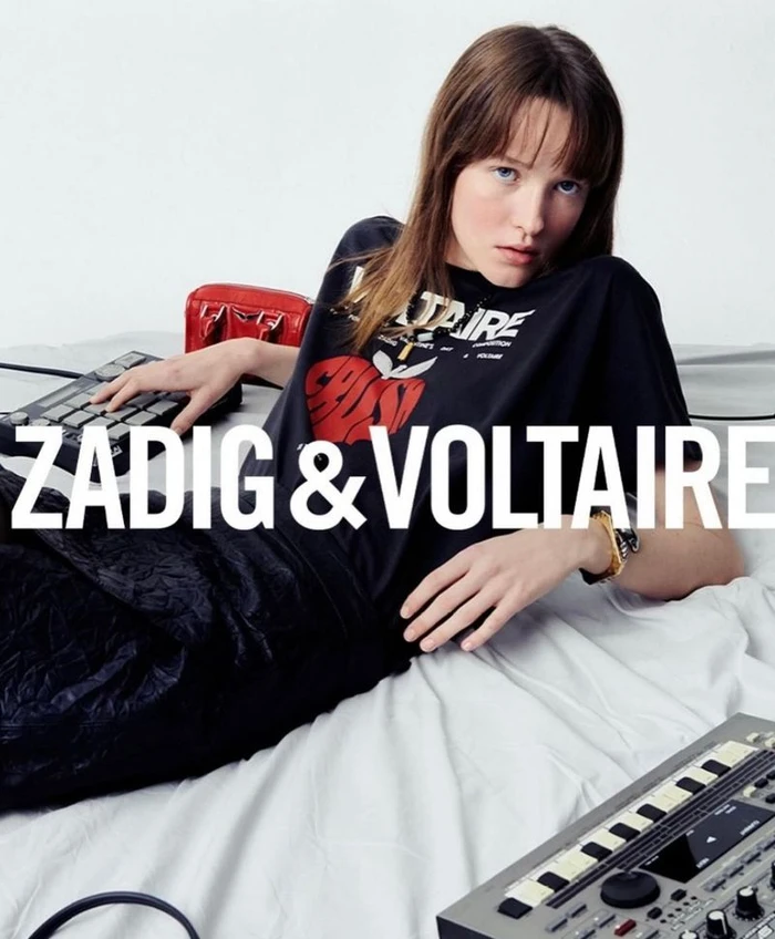 Model lying on white bedsheets wearing black graphic t-shirt with electronic music equipment nearby