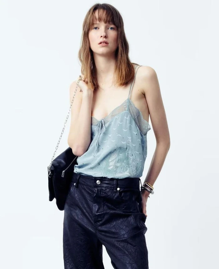 Model wearing a blue camisole and dark trousers holding a black shoulder bag against a white backdrop.