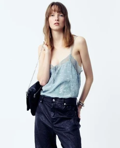 Model wearing a blue camisole and dark trousers holding a black shoulder bag against a white backdrop.