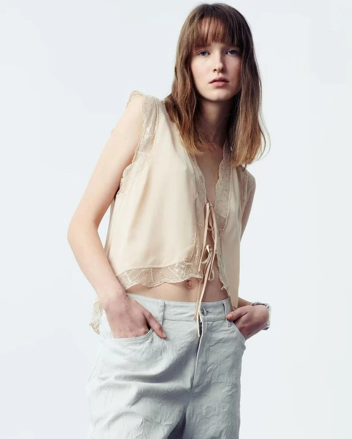 Model wearing a cream lace-trimmed sleeveless top and light trousers against a clean white background