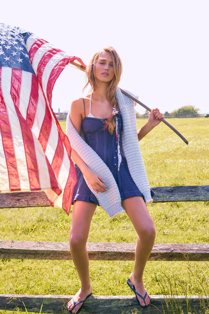 Woman stands by a wooden fence in a grassy field, holding a large American flag and wearing a blue romper and a knitted cardigan.