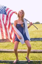 Model holding an American flag and leaning on a wooden fence in a sunny meadow during the LoveShackFancy Hamptons LookBook shoot.