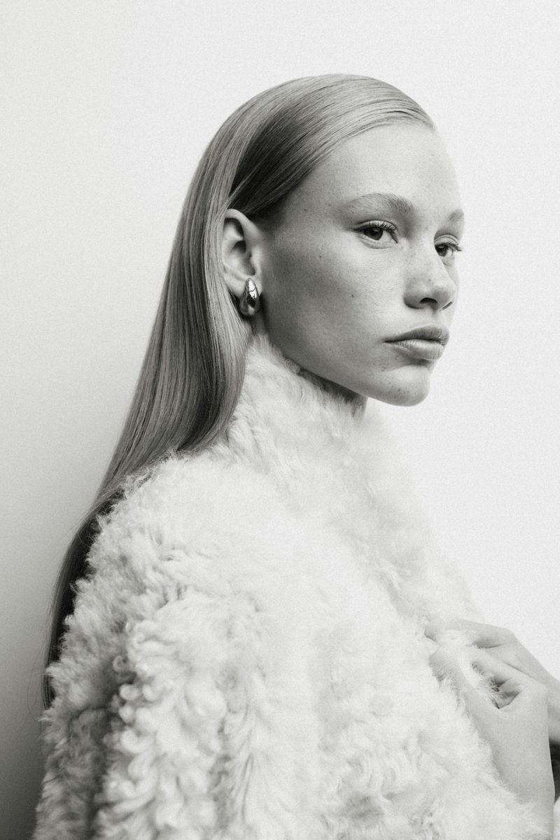 Close-up portrait of a fair-skinned woman with blonde hair wearing silver hoop earrings and a white fuzzy jacket against a white background.