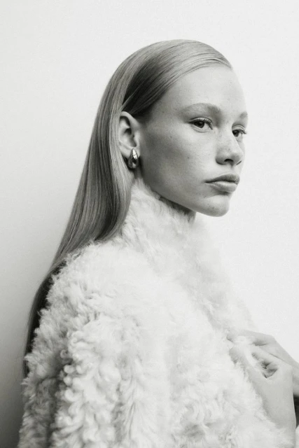 Close-up portrait of a fair-skinned woman with blonde hair wearing silver hoop earrings and a white fuzzy jacket against a white background.