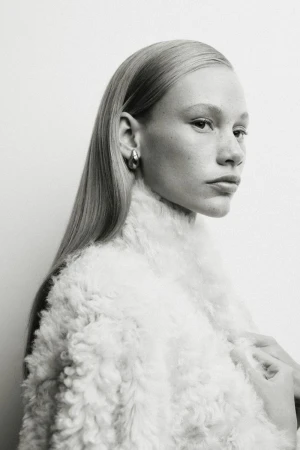 Close-up portrait of a fair-skinned woman with blonde hair wearing silver hoop earrings and a white fuzzy jacket against a white background.