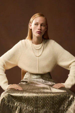Model seated wearing a cream knit sweater, metallic gold skirt, and layered pearl necklaces against a warm brown studio background.