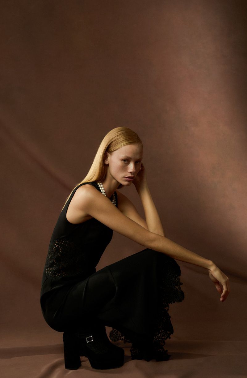 A fair-skinned woman with blonde hair kneels, wearing a black outfit and platform shoes against a solid brown studio backdrop.