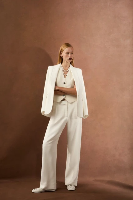 Woman in a cream-colored three-piece suit with arms crossed in a studio setting with a neutral background. She is wearing a pearl necklace.
