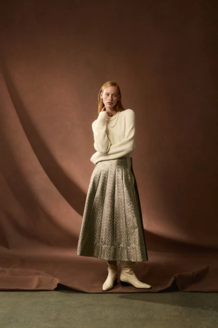 Fashion editorial shot of a female adult model in a cream sweater and a pleated skirt standing against a draped brown backdrop.