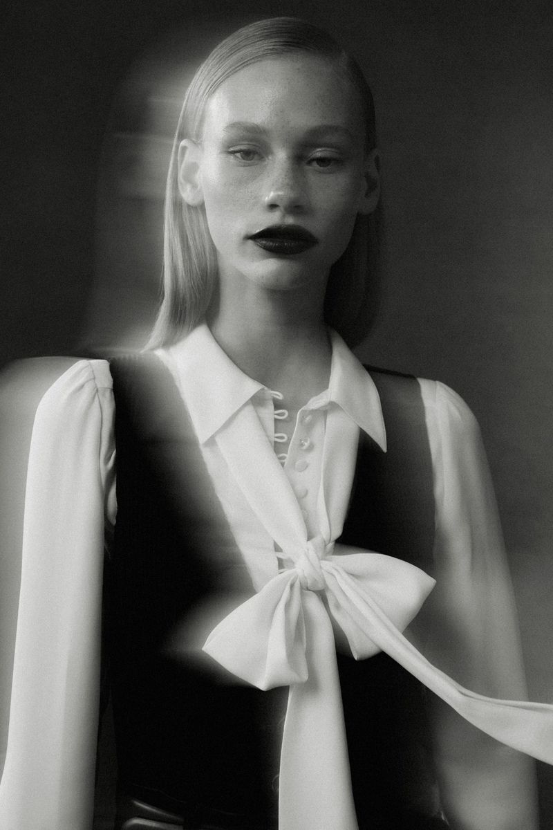 Monochrome fashion portrait of a fair-skinned adult woman with dark lipstick, wearing a white blouse with a large bow and a vest.