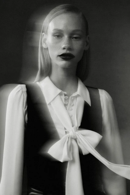 Monochrome fashion portrait of a fair-skinned adult woman with dark lipstick, wearing a white blouse with a large bow and a vest.