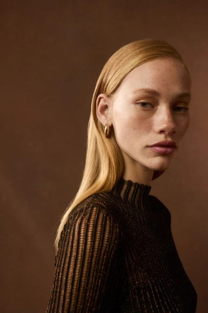 Elegant portrait of a young adult woman with blonde hair, wearing a brown ribbed top, and gold hoop earrings against a brown background.