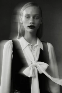 Portrait of a female model wearing a white blouse with a bow and dark vest in a monochrome lookbook shot.
