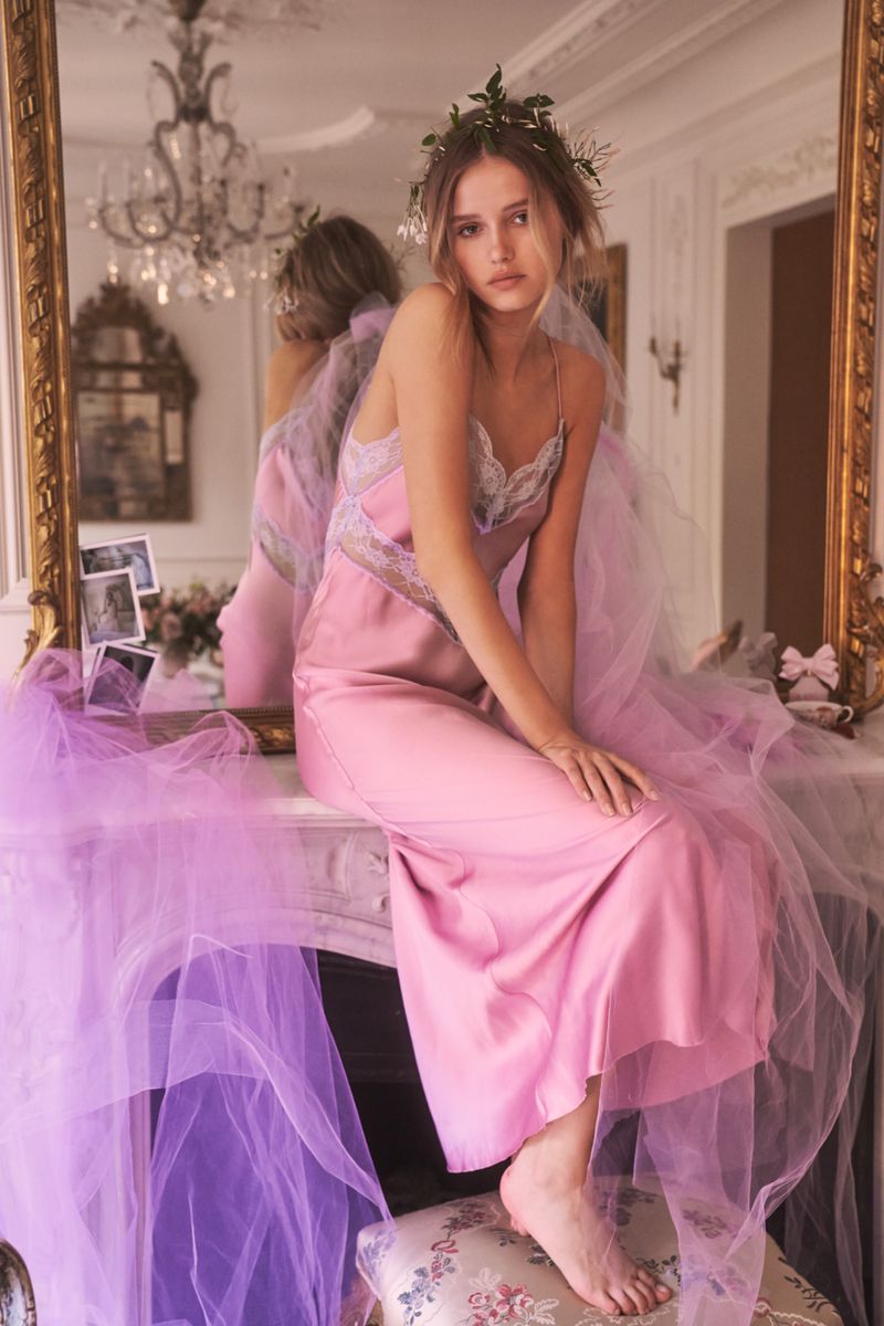 Woman in pink slip dress with lace trim and floral headpiece sits on fireplace mantel with tulle fabric, mirror, and chandelier in background.