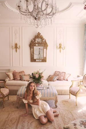 Woman in a cream dress poses on a floral rug in an ornate living room with white couches, decorative mirror, and chandelier.